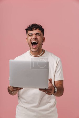 Téléchargez les photos : Jeune homme de plissement enthousiaste beau avec bouche ouverte tenant ordinateur portable et regardant la caméra, tout en se tenant sur fond isolé rose - en image libre de droit