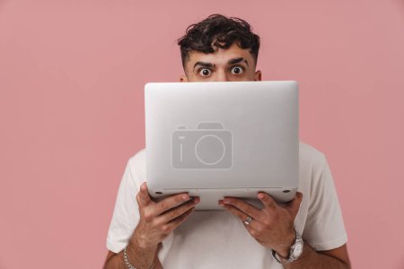 Téléchargez les photos : Jeune bel homme bouclé regardant par derrière un ordinateur portable et regardant la caméra, tout en se tenant debout sur un fond rose isolé - en image libre de droit