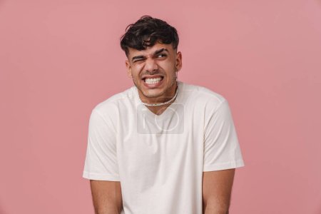 Photo for Young stylish handsome man in white t-shirt and necklace with closed eye and clenched teeth looking aside, while standing over isolated pink background - Royalty Free Image