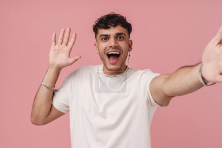 Photo for Selfie of young handsome stylish smiling man with opened mouth in necklace waving and looking at camera , while standing over isolated pink background - Royalty Free Image