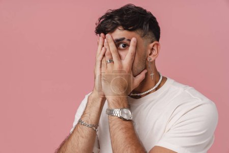 Photo for Young handsome stylish man covering his face and peeping between fingers and looking at camera, while standing over pink isolated background - Royalty Free Image