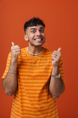 Photo for Young handsome stylish happy smiling man with piercing in necklace showing thumb gesture both hands and looking at camera , while standing over isolated orange background - Royalty Free Image