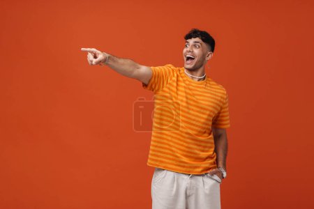 Téléchargez les photos : Portrait de jeune homme élégant heureux et enthousiaste avec la bouche ouverte dans le collier et le t-shirt, pointant et regardant de côté, tout en se tenant la main dans la poche sur fond orange isolé - en image libre de droit