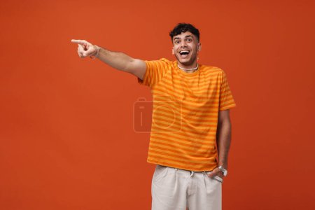 Téléchargez les photos : Portrait de jeune homme élégant et heureux enthousiaste avec la bouche ouverte dans le collier et t-shirt pointant de côté et regardant la caméra, tout en se tenant la main dans la poche sur fond orange isolé - en image libre de droit