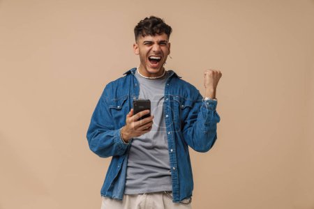 Téléchargez les photos : Jeune homme élégant et enthousiaste tenant le téléphone faisant geste gagnant avec le poing levé et la bouche ouverte, tout en se tenant sur fond brun isolé - en image libre de droit