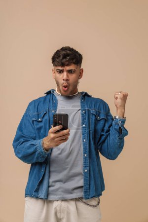 Photo for Young handsome stylish enthusiastic man holding phone doing winner gesture with raised fist and opened mouth , while standing over isolated brown background - Royalty Free Image
