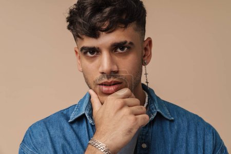 Photo for Portrait of young handsome stylish thoughtful man with piercing in necklace and jeans shirt touching his chin and looking at camera, while standing over isolated brown background - Royalty Free Image