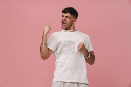 Foto de Joven hombre guapo y elegante en camiseta blanca con los ojos cerrados, la boca abierta y las manos levantadas bailando sobre un fondo rosa aislado - Imagen libre de derechos
