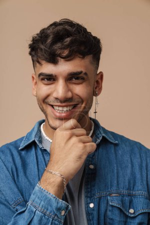 Photo for Portrait of young handsome stylish smiling happy man with piercing in necklace and jeans shirt touching his chin and looking at camera, while standing over isolated brown background - Royalty Free Image