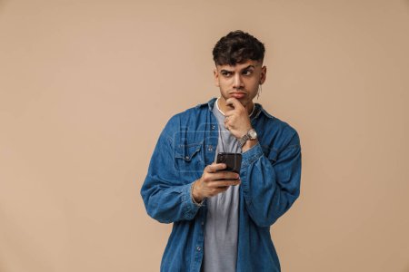 Photo for Brunette young man wearing shirt frowning and using cellphone isolated over beige background - Royalty Free Image