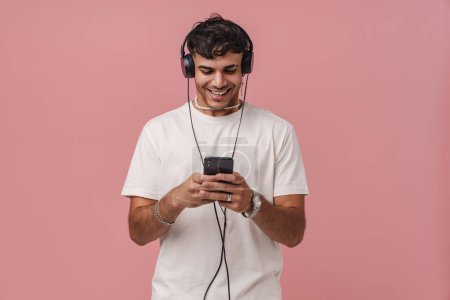Téléchargez les photos : Brunette jeune homme dans un casque souriant tout en utilisant un téléphone mobile isolé sur fond rose - en image libre de droit
