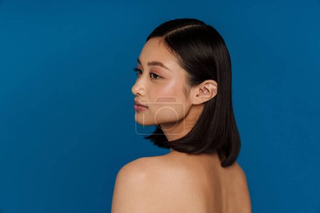 Téléchargez les photos : Portrait de jeune belle belle femme asiatique calme regardant de côté, tout en se tenant avec son dos sur fond bleu isolé - en image libre de droit