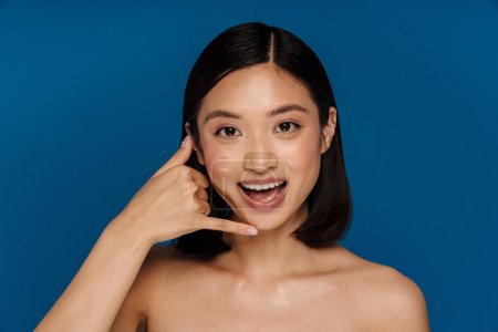 Téléchargez les photos : Jeune belle femme asiatique souriante heureuse avec bouche ouverte faisant geste de téléphone en regardant la caméra, tout en se tenant debout sur fond bleu isolé - en image libre de droit