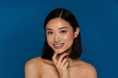Téléchargez les photos : Jeune belle heureuse souriante asiatique femme toucher son menton en regardant la caméra, tout en se tenant sur fond bleu isolé - en image libre de droit