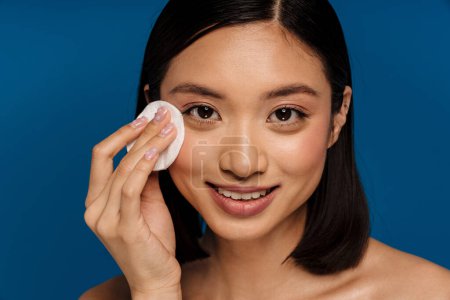 Téléchargez les photos : Portrat de jeune belle femme asiatique souriante essuyant son visage avec du coton et regardant la caméra, tout en se tenant sur fond bleu isolé - en image libre de droit