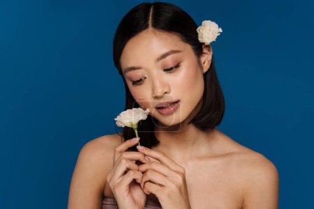 Photo for Young beautiful asian woman with opened mouth with flower on ear holding white flower and looking on it, while standing over isolated blue background - Royalty Free Image