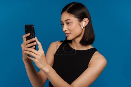 Foto de Asiática joven mujer en auriculares sonriendo mientras se utiliza el teléfono móvil aislado sobre fondo azul - Imagen libre de derechos