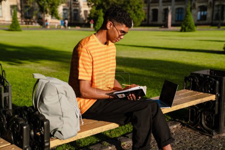 Photo for Young african man student in casual wear writing down notes while sitting on bench in university campus outdoors - Royalty Free Image