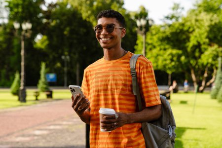 Photo for Young black brunette man wearing sunglasses walking outdoors with cup of coffee and cellphone - Royalty Free Image