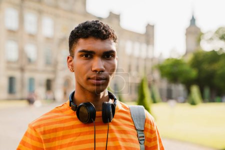 Téléchargez les photos : Jeune homme brune noire avec perçage et écouteurs marchant par l'université en plein air - en image libre de droit