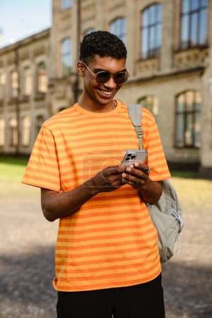 Téléchargez les photos : Jeune homme brune noire portant des lunettes de soleil marchant à l'extérieur avec téléphone portable - en image libre de droit
