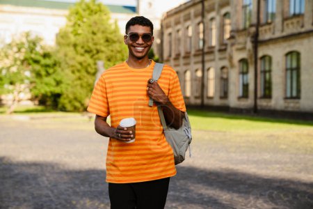 Photo for Young black brunette man wearing piercing and sunglasses walking by college with cup of coffee - Royalty Free Image