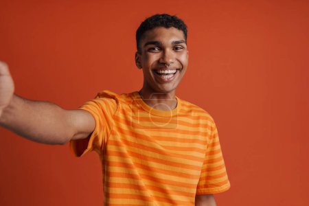Foto de Autorretrato de joven guapo sonriente hombre africano feliz en camiseta mirando a la cámara, mientras está de pie sobre un fondo naranja aislado - Imagen libre de derechos