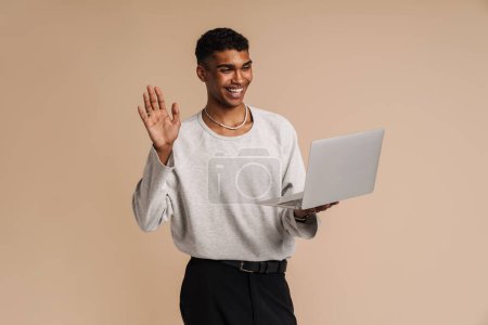 Téléchargez les photos : Jeune homme noir agitant la main tout en travaillant avec un ordinateur portable isolé sur fond beige - en image libre de droit