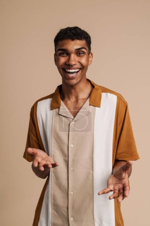 Téléchargez les photos : Jeune homme noir avec des gestes perçants tout en riant à la caméra isolé sur fond beige - en image libre de droit