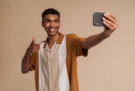 Téléchargez les photos : Jeune homme africain beau et souriant prenant selfie avec téléphone et montrant le geste du pouce, tout en se tenant debout sur fond beige isolé - en image libre de droit