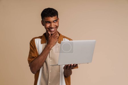 Téléchargez les photos : Jeune homme africain souriant avec percing tenant un ordinateur portable, touchant son menton et regardant de côté, tout en se tenant sur fond beige isolé - en image libre de droit