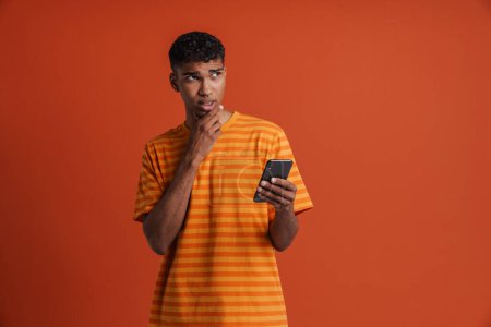 Téléchargez les photos : Jeune homme africain attentionné et beau avec un piercing tenant son téléphone, regardant de côté et touchant son menton, debout sur un fond orange isolé - en image libre de droit