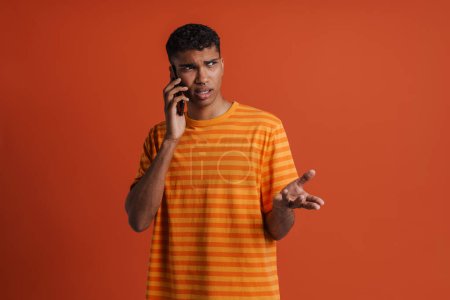 Foto de Joven guapo hombre africano serio con piercing hablando por teléfono y mirando a un lado, mientras está de pie sobre un fondo naranja aislado - Imagen libre de derechos