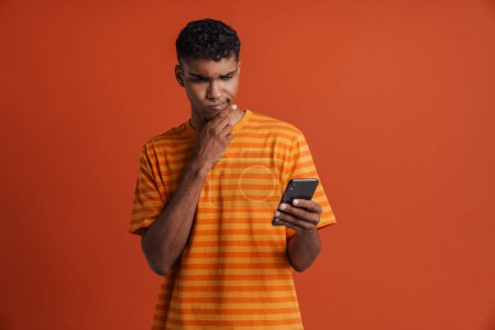 Téléchargez les photos : Jeune bel homme africain réfléchi avec percing tenant son téléphone en le regardant et en touchant son menton, tout en se tenant debout sur un fond orange isolé - en image libre de droit