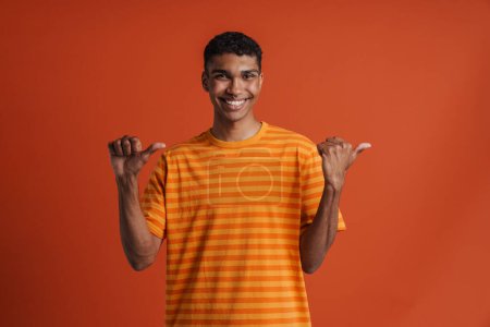 Photo for Young handsome smiling happy african man with piercing pointing aside with both thumbs and looking at camera, while standing over isolated orange background - Royalty Free Image