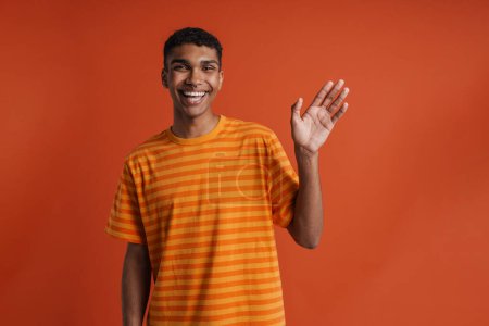 Foto de Joven guapo sonriente hombre africano feliz con piercing saludando y mirando a la cámara, mientras que de pie sobre fondo naranja aislado - Imagen libre de derechos