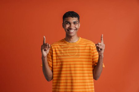 Téléchargez les photos : Jeune homme africain heureux et souriant avec un piercing pointant vers le haut avec les deux mains et regardant la caméra, tout en se tenant debout sur un fond orange isolé - en image libre de droit