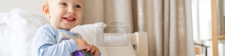 Foto de Feliz bebé rubio jugando con juguetes mientras está sentado en la cama en casa - Imagen libre de derechos