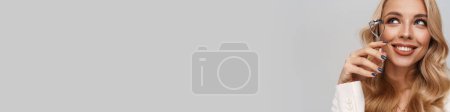 Téléchargez les photos : Joyeux magnifique femme souriant tout en posant avec friseur de cils isolé sur fond gris - en image libre de droit