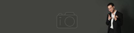 Téléchargez les photos : Unshaven brunette homme souriant et pointant du doigt à la caméra isolé sur fond noir - en image libre de droit