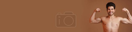 Téléchargez les photos : Homme heureux torse nu souriant et montrant ses biceps à la caméra isolé sur fond beige - en image libre de droit