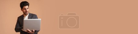 Téléchargez les photos : Jeune homme sérieux en veste debout tout en travaillant avec un ordinateur portable isolé sur fond beige - en image libre de droit