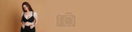 Téléchargez les photos : Souriant jeune femme taille plus portant soutien-gorge posant sur fond mural beige - en image libre de droit