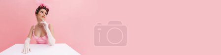 Téléchargez les photos : Jeune femme portant des gants regardant de côté tout en étant assis à la table isolé sur fond rose - en image libre de droit