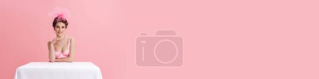Téléchargez les photos : Jeune femme portant des bigoudis souriant à la caméra tout en étant assis à la table isolé sur fond rose - en image libre de droit