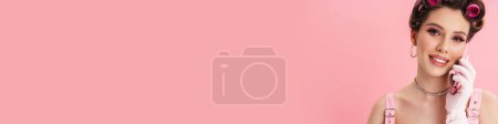 Téléchargez les photos : Jeune femme avec des bigoudis souriant tout en parlant sur smartphone isolé sur fond rose - en image libre de droit