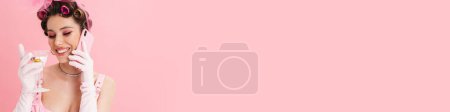 Téléchargez les photos : Jeune femme avec des bigoudis boire cocktail tout en parlant sur smartphone isolé sur fond rose - en image libre de droit