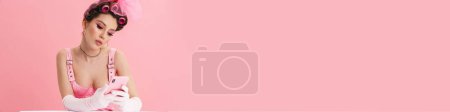 Téléchargez les photos : Jeune femme avec des bigoudis posant et utilisant un téléphone portable isolé sur fond rose - en image libre de droit