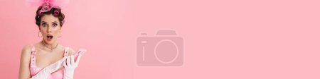 Téléchargez les photos : Femme surprise avec des bigoudis s'écriant tout en utilisant un téléphone mobile isolé sur fond rose - en image libre de droit