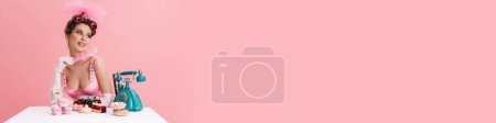 Téléchargez les photos : Jeune femme portant des bigoudis tenant plume tout en étant assis à la table isolé sur fond rose - en image libre de droit